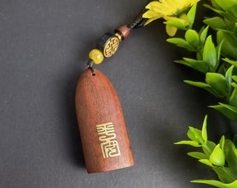 May include: A wooden pendant with gold-colored Chinese characters, attached to a black cord with decorative beads and a yellow flower. The pendant is a rich brown color and has a smooth finish.