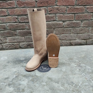 May include: A pair of beige leather knee-high boots with a pointed toe and a low heel. The boots are shown on a concrete surface with a brick wall in the background.