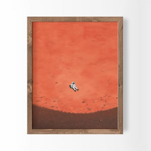 Peut inclure: Une impression d'art encadrée représentant un petit astronaute en combinaison blanche allongé sur une vaste surface texturée orange-rouge. La partie inférieure montre une ligne d'horizon courbe brun foncé contrastant avec le paysage orange.