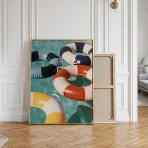 May include: A rectangular wall art print featuring three inflatable pool rings floating on turquoise water. One ring is yellow and white, one is orange and white, and one is dark blue and white. Each ring has a small white handle attached to the side.
