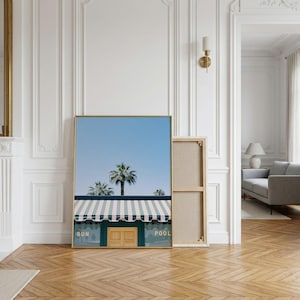 May include: A rectangular wall art print featuring a clear blue sky above palm trees and a building with a green facade, a striped awning, and a central wooden door. The text SUN and POOL is printed in white on the green wall sections.