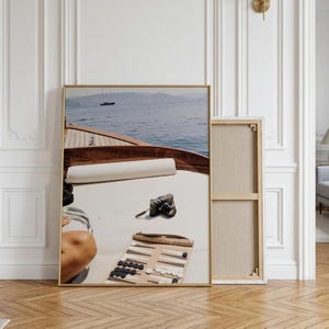 May include: A framed canvas print depicting a view from the deck of a wooden boat looking out over blue water toward distant mountains. On the white deck surface sits a black camera and a tan backgammon board with black and white game pieces.
