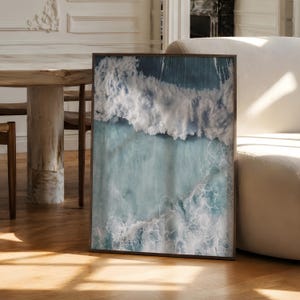 May include: A rectangular framed wall art print featuring an aerial view of ocean waves crashing. The image displays deep blue and turquoise water with white sea foam patterns across the surface, enclosed in a thin, dark brown wooden frame.