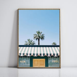 Op de afbeelding: Ingelijste print met een zonnige scène met palmbomen tegen een helderblauwe lucht. De afbeelding bevat een gebouw met een gestreepte luifel en de woorden "SUN" en "POOL".