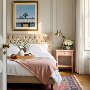 May include: A bedroom featuring a beige tufted headboard, a white bed with a pink throw blanket, and a woven tray holding a white vase with white flowers. A pink nightstand with a brass lamp and white flowers sits beside the bed.