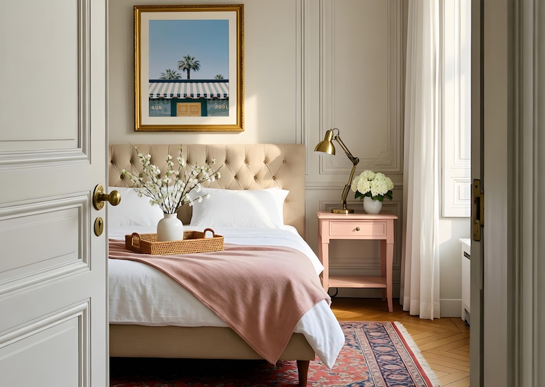 May include: A bedroom featuring a beige tufted headboard, a white bed with a pink throw blanket, and a woven tray holding a white vase with white flowers. A pink nightstand with a brass lamp and white flowers sits beside the bed.
