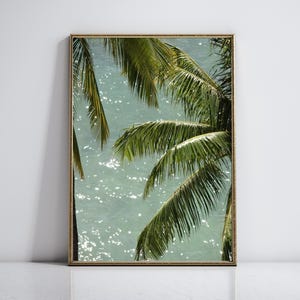 Op de afbeelding: Een ingelijste kunstdruk met een tropische scène. De afbeelding toont levendige groene palmbladeren tegen een achtergrond van glinsterend turquoise water. Het kunstwerk is ingesloten in een goudkleurige lijst, wat een vleugje elegantie geeft.