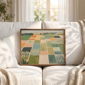 May include: A rectangular wall art print in a dark wood frame featuring an aerial landscape painting of agricultural fields. The composition displays a grid of rectangular plots in various shades of green, yellow, orange, and beige, depicting crops.