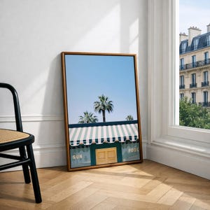 May include: A framed vertical wall art print featuring a light blue sky, palm trees, and a building with a green and white striped awning. The building facade includes a wooden door and the text SUN and POOL printed in gold lettering.