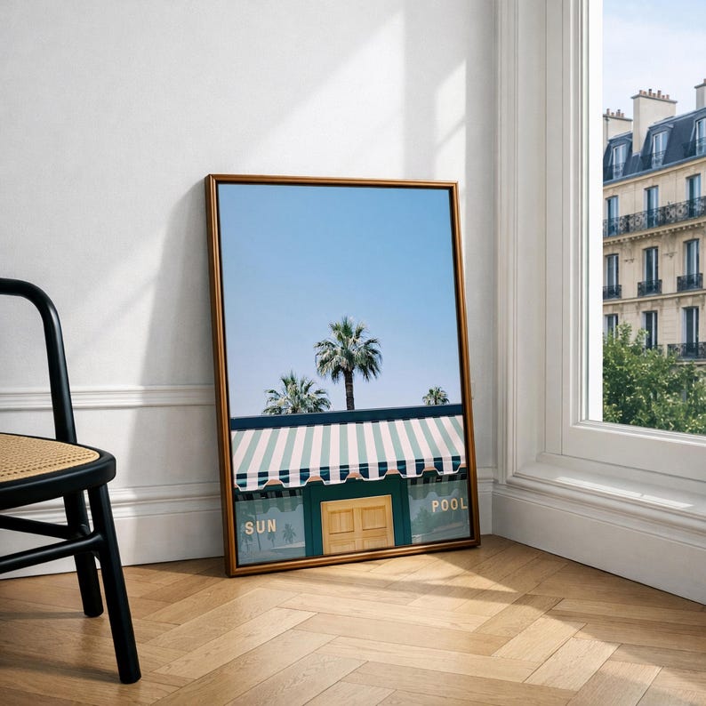 May include: A framed vertical wall art print featuring a light blue sky, palm trees, and a building with a green and white striped awning. The building facade includes a wooden door and the text SUN and POOL printed in gold lettering.