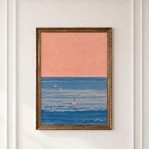 May include: A framed wall art print featuring a textured painting of a calm blue ocean under a solid peach-colored sky. Three small figures are visible on paddleboards in the water, rendered in a simple, minimalist style with visible brushstrokes.