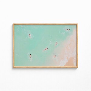 May include: A framed horizontal art print featuring an aerial view of a beach with turquoise water and light sand. Several small figures are swimming in the ocean, while white sea foam washes onto the shore in this minimalist coastal scene.