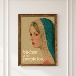 May include: A framed wall art print featuring a portrait of a person with a teal head covering and lace trim. The person has light skin and red lips. The text "bitches are people too." is printed in white in the lower left corner of the print.