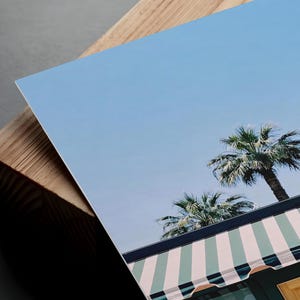 May include: A rectangular photographic print featuring a clear blue sky above two palm trees and a building with a green and white striped awning. The print is angled and rests against a thick, light-colored wooden block with visible grain.
