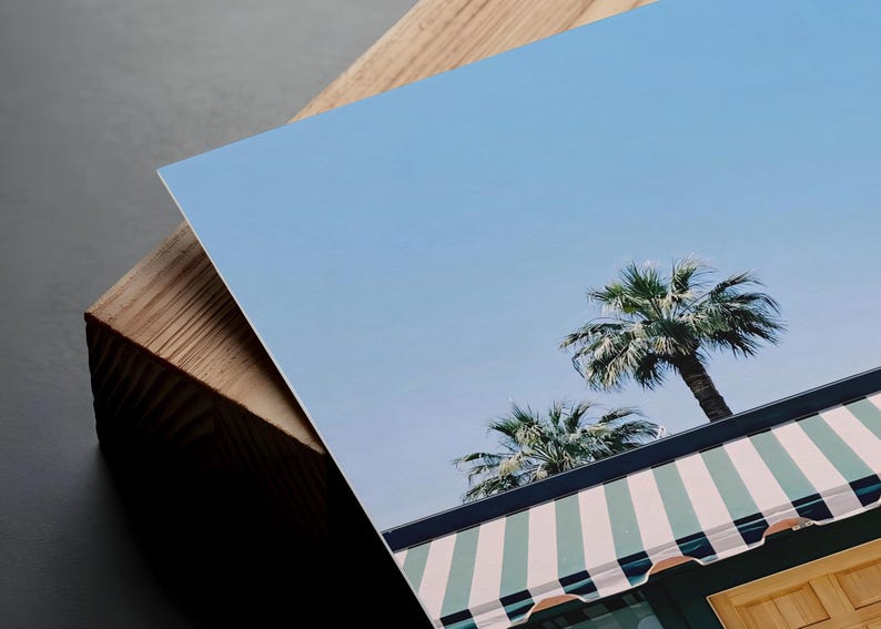 May include: A rectangular photographic print featuring a clear blue sky above two palm trees and a building with a green and white striped awning. The print is angled and rests against a thick, light-colored wooden block with visible grain.
