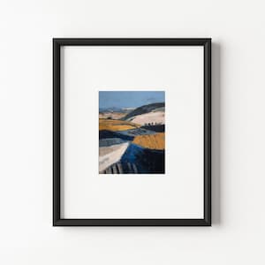 Puede incluir: Un cuadro de paisaje enmarcado que muestra un terreno ondulado con parches de amarillo dorado, azul profundo y blanco. La obra está dentro de un marco negro sencillo con un paspartú blanco ancho, mostrando colinas con textura.