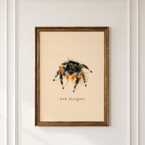 May include: A framed wall art print featuring a detailed illustration of a black jumping spider with orange accents on its legs. The spider is centered on a light beige background with the text "web designer" printed in small, dark letters below it.