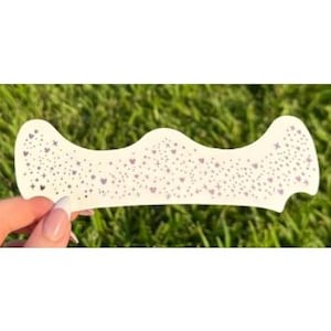 May include: A white nail art stencil with a wavy top edge and a decorative pattern of small hearts and dots. The stencil is held against a background of green grass.