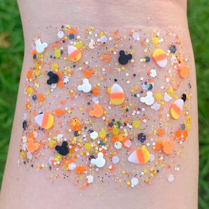 Puede incluir: Una superficie transparente y brillante con una mezcla de confeti naranja, amarillo, blanco y negro. El diseño incluye formas de caramelos de maíz, siluetas de Mickey Mouse y pequeñas piezas hexagonales. El efecto general es festivo y colorido.