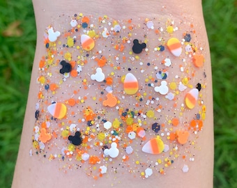 May include: A clear, glittery surface with a mix of orange, yellow, white, and black confetti. The design includes candy corn shapes, Mickey Mouse silhouettes, and small hexagonal pieces. The overall effect is festive and colorful.