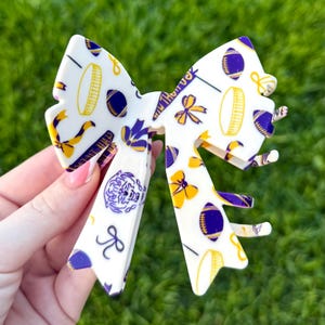 May include: White and purple bow hair clip with a repeating pattern of footballs, flowers, and the text "Geaux Tigers". The bow is held in front of a green grassy background.