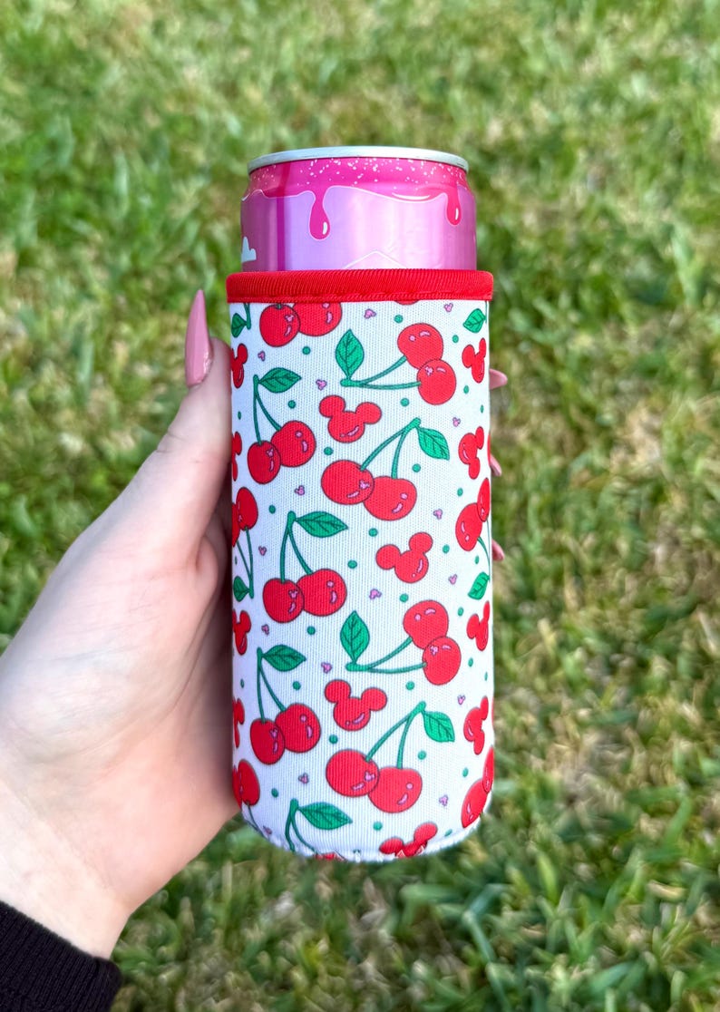 May include: A white can cooler with a red trim and a pattern of red cherries and green leaves. The cooler is holding a pink can. The background is green grass.