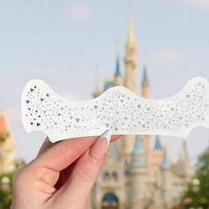 May include: A white, curved decorative piece with a pattern of small stars and Mickey Mouse silhouettes. The piece is held up against a blurred background of a castle and buildings, suggesting a theme park setting.