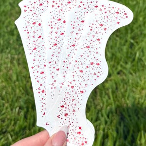 May include: A stack of white, decorative stickers with a unique shape. Each sticker is adorned with a repeating pattern of red stars, dots, and other small shapes. The stickers are held against a backdrop of green grass.