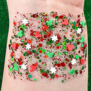 May include: A clear, glittery gel applied to the skin, featuring red, green, and white glitter in various shapes and sizes. The glitter includes Mickey Mouse silhouettes and snowflake shapes, creating a festive appearance.