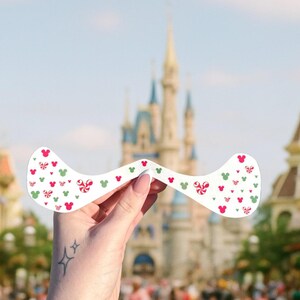 May include: A white decorative item with a curved shape, adorned with red Mickey Mouse silhouettes, green Mickey heads, and red and white candy cane designs. The background features a blurred view of a castle and a sunny sky.