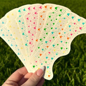 May include: A fan-shaped collection of cream-colored stickers, each adorned with colorful heart-shaped and dot patterns. The stickers are held against a backdrop of green grass, showcasing a vibrant and playful design.