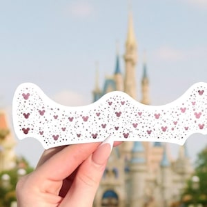 May include: A white decorative item with a wavy top edge, adorned with small, dark pink Mickey Mouse head shapes and dots. The background features a blurred view of a castle and buildings, suggesting a theme park setting.