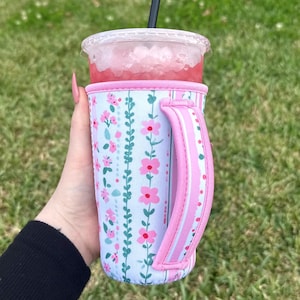 以下が含まれることがあります： ピンクのハンドルとトリムが付いた花柄のドリンクスリーブ。ピンク色の飲み物と氷が入った透明なプラスチックカップが入っています。スリーブには、薄い青色の背景にピンクの花と緑のつるが描かれています。
