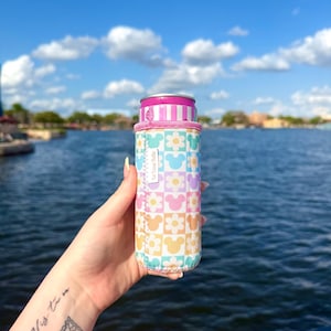 Pode incluir: Um refrigerador de latas colorido com um padrão floral e Mickey Mouse em cores pastel. O refrigerador tem um topo listrado rosa e branco e é segurado por uma mão contra um fundo de água e céu azul.