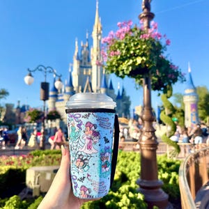 Könnte beinhalten: Ein hellblauer isolierter Becher mit Disney-Figuren in einem skurrilen Design. Der Becher hat einen schwarzen Griff und einen transparenten Deckel. Im Hintergrund sind ein Schloss und ein sonniger Tag zu sehen.