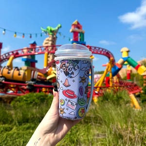 K&ouml;nnte beinhalten: Ein hellvioletter Reisebecher mit einem durchsichtigen Deckel und Griff, der ein farbenfrohes Toy Story-Design aufweist. Das Design enth&auml;lt Bilder von Woody, Buzz Lightyear und anderen Charakteren. Der Hintergrund zeigt einen Vergn&uuml;gungspark mit einer Achterbahn.