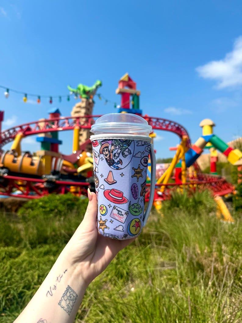 K&ouml;nnte beinhalten: Ein hellvioletter Reisebecher mit einem durchsichtigen Deckel und Griff, der ein farbenfrohes Toy Story-Design aufweist. Das Design enth&auml;lt Bilder von Woody, Buzz Lightyear und anderen Charakteren. Der Hintergrund zeigt einen Vergn&uuml;gungspark mit einer Achterbahn.