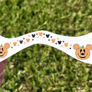 May include: A white, curved Halloween-themed decoration featuring orange Mickey Mouse pumpkin faces and black Mickey Mouse silhouettes. The decoration is held against a backdrop of green grass.