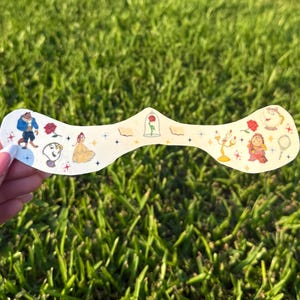 May include: A decorative, off-white, curved sticker featuring illustrations of characters and objects from the animated film "Beauty and the Beast." The sticker includes images of Belle, the Beast, Lumiere, Mrs. Potts, and the enchanted rose. The sticker is held by a person.