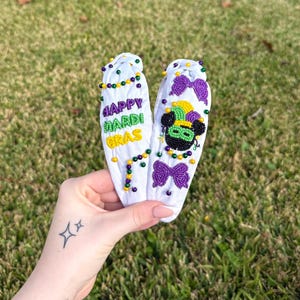 May include: Two white headbands with Mardi Gras designs. One headband has the text "HAPPY MARDI GRAS" in purple, green, and yellow beads. The other headband features a Mickey Mouse-inspired design with a green and black mask, purple bows, and yellow and green beads.