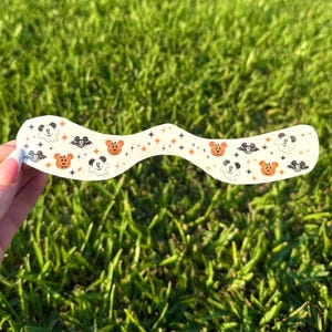 May include: A white, curved sticker with a Halloween theme. The sticker features orange jack-o'-lanterns, black bats, Mickey Mouse, and ghosts. The design is accented with orange and black stars. The sticker is held against a background of green grass.
