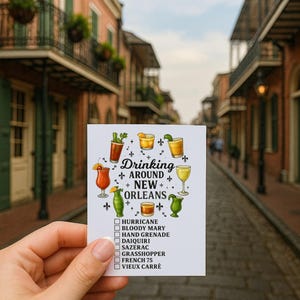 Può includere: Una carta bianca con il testo "Drinking Around New Orleans" e illustrazioni di cocktail. La carta elenca vari cocktail di New Orleans, tra cui Hurricane, Bloody Mary e Sazerac. La carta è tenuta davanti a una scena stradale sfocata.