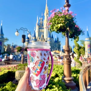 Könnte beinhalten: Ein weißer Becher mit rosa Griff und Rand, verziert mit einem Alice im Wunderland-Design. Der Becher wird vor einem Schloss und blauem Himmel gehalten. Das Design umfasst Charaktere und Elemente aus der klassischen Geschichte.