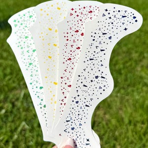 May include: A hand holds five white nail art stencils with a variety of colorful designs. Each stencil features a different color of embellishments, including green, yellow, red, and blue, with stars, dots, and other shapes.