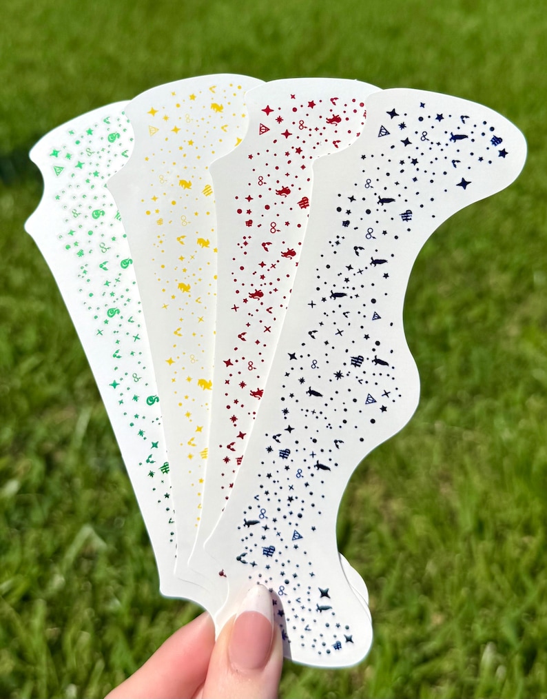 May include: A hand holds five white nail art stencils with a variety of colorful designs. Each stencil features a different color of embellishments, including green, yellow, red, and blue, with stars, dots, and other shapes.