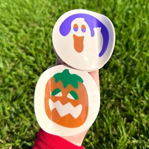 May include: Two small, round Halloween-themed dishes. One features a purple ghost with an orange mouth, and the other has an orange pumpkin with a green stem and a white, toothy grin. The dishes are set against a backdrop of green grass.