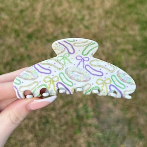 May include: White hair claw clip with a Mardi Gras theme. The clip features a pattern of purple, green, and gold beads, bows, and donuts. The claw clip is held by a hand against a blurred green background.