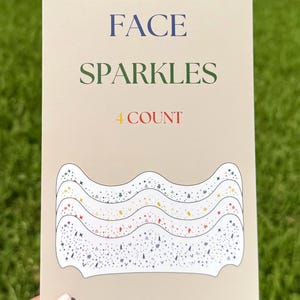 May include: A beige card with the words "FACE SPARKLES 4 COUNT" in blue, green, and red. The card features a wavy design with various colored sparkles and star shapes, perfect for face decoration.