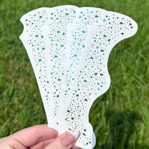 May include: A stack of white, curved stencils with a green celestial pattern of stars, dots, and symbols. The stencils are held against a backdrop of green grass, suggesting an outdoor setting. The stencils are likely for crafting or decorative purposes.