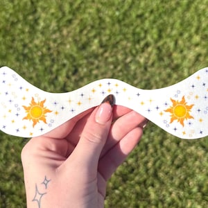 Peut inclure: Un autocollant décoratif blanc, aux bords ondulés, représentant un soleil jaune avec des rayons orange et des étoiles bleues et jaunes. L'autocollant est tenu sur un fond herbeux vert.